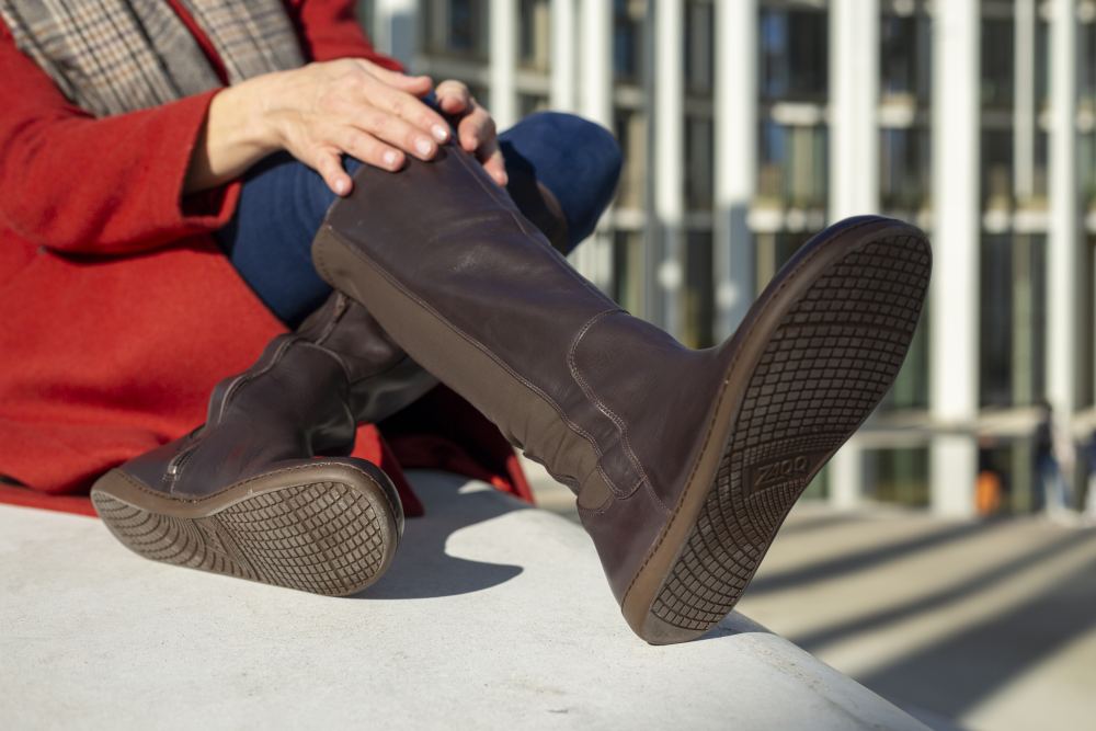 A person in a red coat, blue jeans and knee-high boots QATCH Nappa Brown by ZAQQ sits outside with her feet on a surface. A modern building with vertical white lines can be seen in the background.