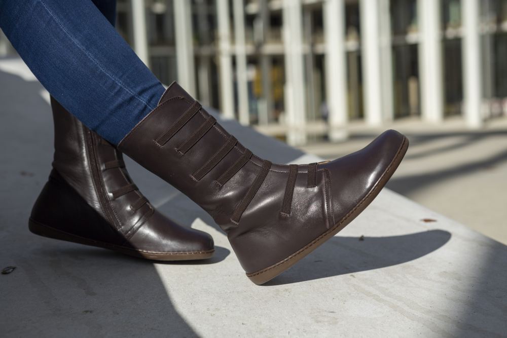 A person wearing ZAQQ's ATTRAQT Stretch Nappa Brown boots and blue jeans sits on a concrete surface. A modern glass facade of a building can be seen in the background.