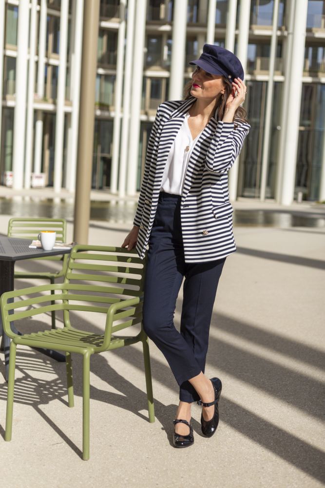 A woman wearing a navy blue cap and striped blazer stands next to a green table and chairs outdoors. She is wearing a white blouse, navy blue pants and TWIST CASEMIRO shoes ZAQQ with flexible soles. Sunlight casts strong shadows on the ground. There is a cup on the table next to her.