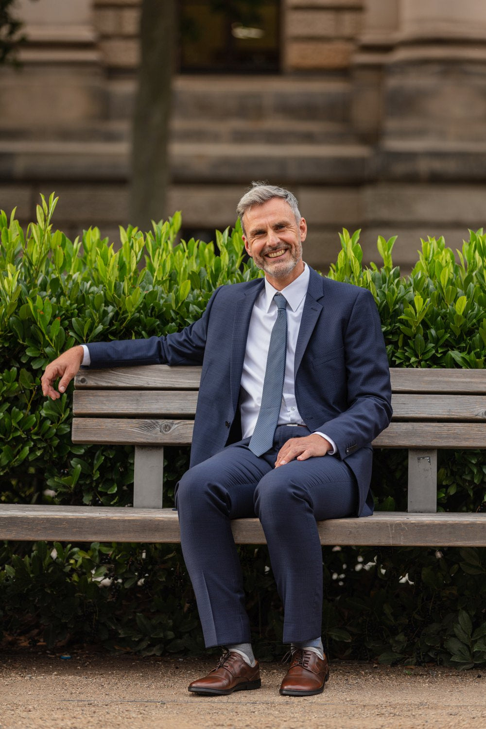 A man wearing a navy blue suit and tie sits on a wooden park bench and smiles carefree. Its polished BRIQ Antique Brown from ZAQQ peeks out from under his pants as he sunbathes in the greenery. A blurred building in the background highlights this calm yet professional moment.