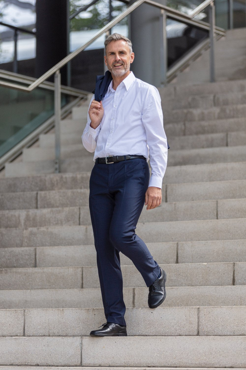 A man with gray hair and a mustache confidently walks down an outdoor staircase, displaying timeless elegance. He wears a white shirt and blue pants and carries his jacket over his shoulder. It therefore embodies the essence of a BRIQ Black design ZAQQ.