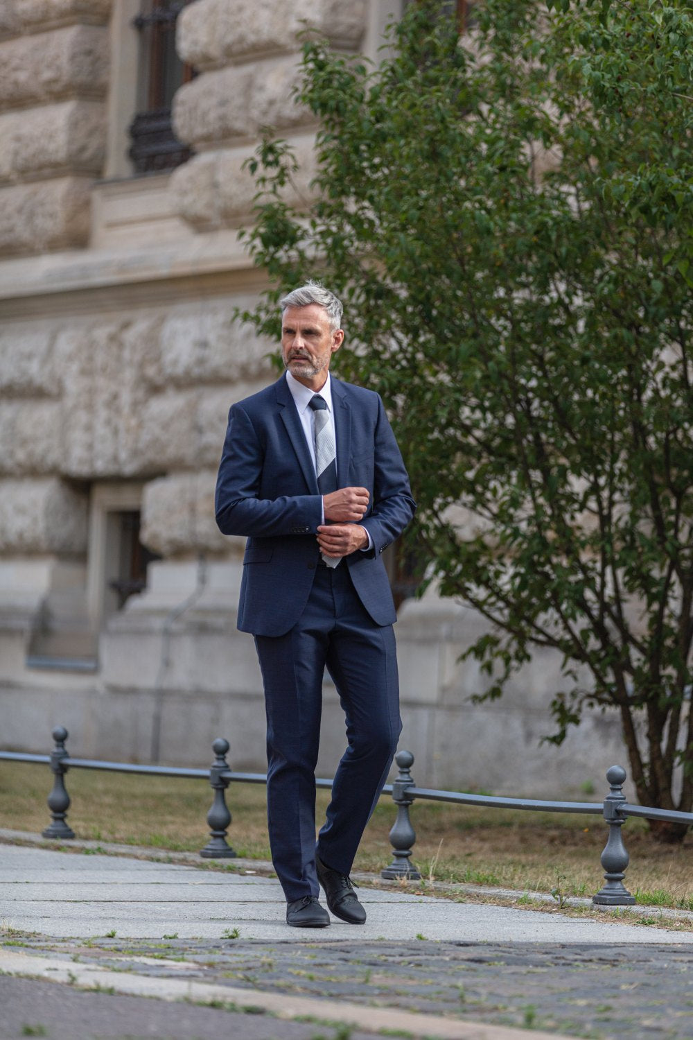 A man in a blue suit and tie looking at the shoes ZAQQ Wearing BRIQ Brogue Vegan Black, she walks along a sidewalk next to a stone building. He has gray hair and a beard. A large bushy tree and metal railing create the perfect backdrop for his stylish path.