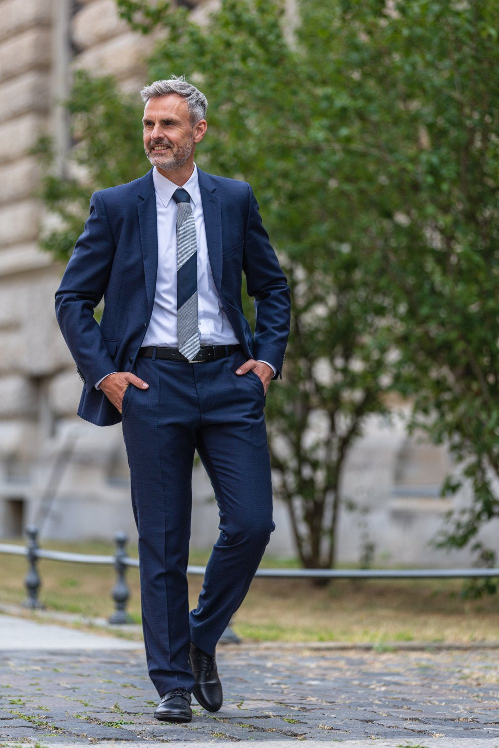 A man with gray hair and a beard walks confidently outside wearing elegant BRIQ Captoe Black shoes from ZAQQ. He combines it with a dark blue suit, a white shirt and a striped tie. With one hand in his pocket, he strolls past a building and trees.