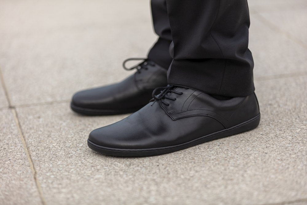 Close-up of a person wearing black PEAQ shoes ZAQQ wears, with elegant leather and comfortable soles, combined with black trousers on a concrete surface. The shoes have laces and a smooth surface and perfectly complement formal wear.
