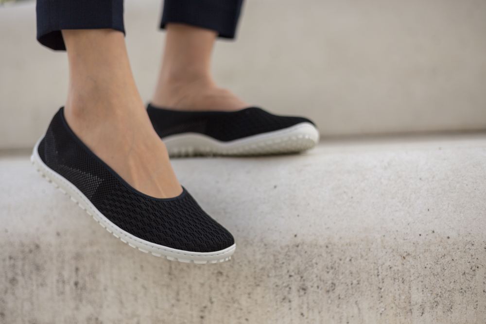 One person is wearing SUQAR Black by ZAQQ, with a smooth mesh-like texture and white sneaker sole, and stands on concrete steps. The focus artfully captures her feet in these slippers as well as part of her lower legs in dark trousers, combining comfort with a subtly feminine look.