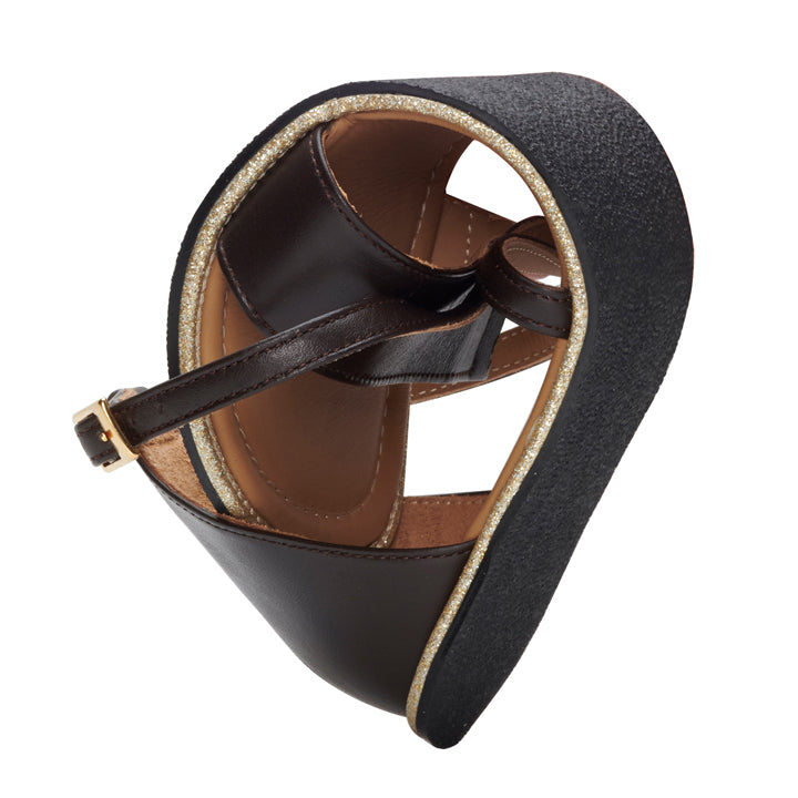 A close-up of a dark brown QEEP sandal from ZAQQ with a shiny gold rim. Crafted from smooth nappa leather, the sandal is folded to reveal its flexible sole that ensures excellent comfort. The dark brown strap features a small gold buckle that contrasts beautifully with the detailed texture of the sole.