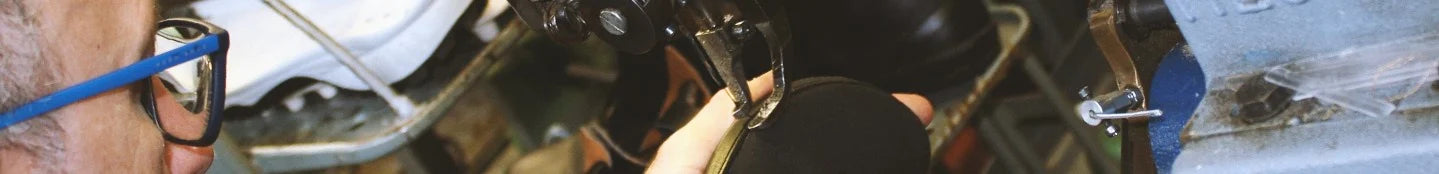 Eine Person mit einer blau umrandeten Brille arbeitet mit einem Werkzeug an einem schwarzen Schuh, vermutlich in einer Werkstatt. Der Hintergrund ist voller unscharfer Objekte und Geräte, was auf eine geschäftige, überfüllte Umgebung hindeutet. Der Fokus liegt auf der Hand und dem Schuh, wodurch der handwerkliche oder Reparaturprozess hervorgehoben wird.