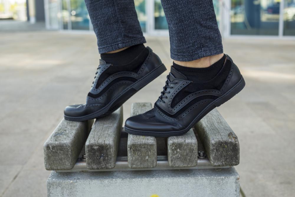 A person in the ZAQQ BRIQ Brogue Deep Black shoes stands confidently on a wooden bench outdoors. These shoes feature decorative perforations that perfectly complement the dark tailored trousers against a softly blurred background.