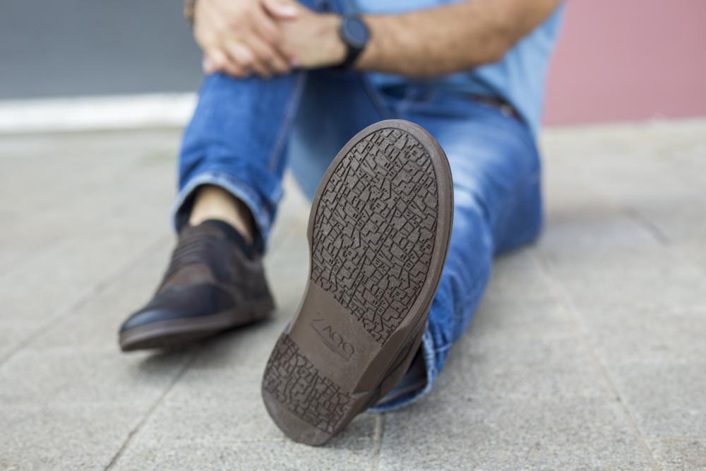 A person sits cross-legged on the floor wearing jeans and a pair ZAQQ BRIQ Brogue Tonka shoes. The focus is on the sole of a shoe, flaunting its intricate patterns. Her hands are folded on her knee, against a blurred background of sidewalk and wall.