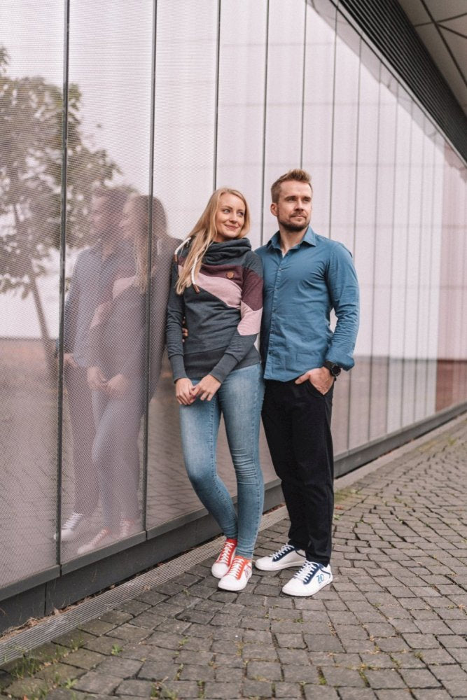 A man and a woman stand leaning close to each other against a modern glass building. The woman, dressed in jeans and a colorful sweater, wears red sneakers, while the man wears dark pants and CEDRIQ Blue barefoot shoes ZAQQ carries. Her reflection shimmers on the glass.
