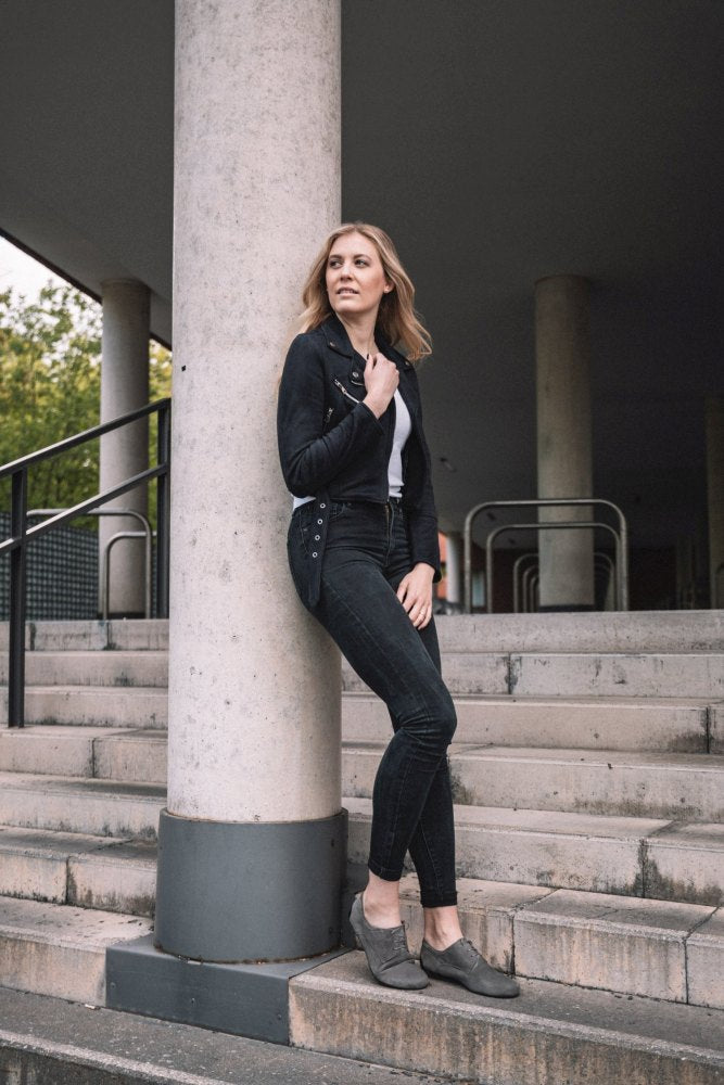 A woman in a gray IQON velor jacket and jeans from ZAQQ leans against a pillar on the steps, perfectly embodying the business model style. With one hand in her pocket and the other in her hair, she exudes confidence. The background of columns and subtle greenery completes the scene.