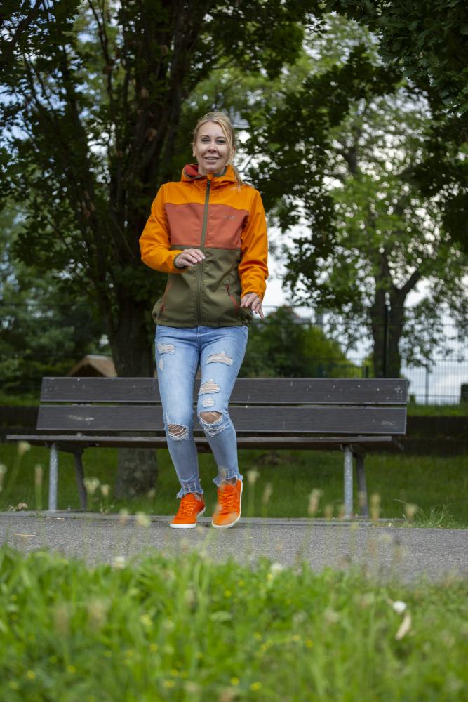 A woman wearing an orange-green jacket and ripped jeans walks along a path in the park, enjoying the barefoot feeling of her bright orange LIQE shoes ZAQQ convey. Smiling, she walks past a wooden bench and green trees under a cloudy sky.
