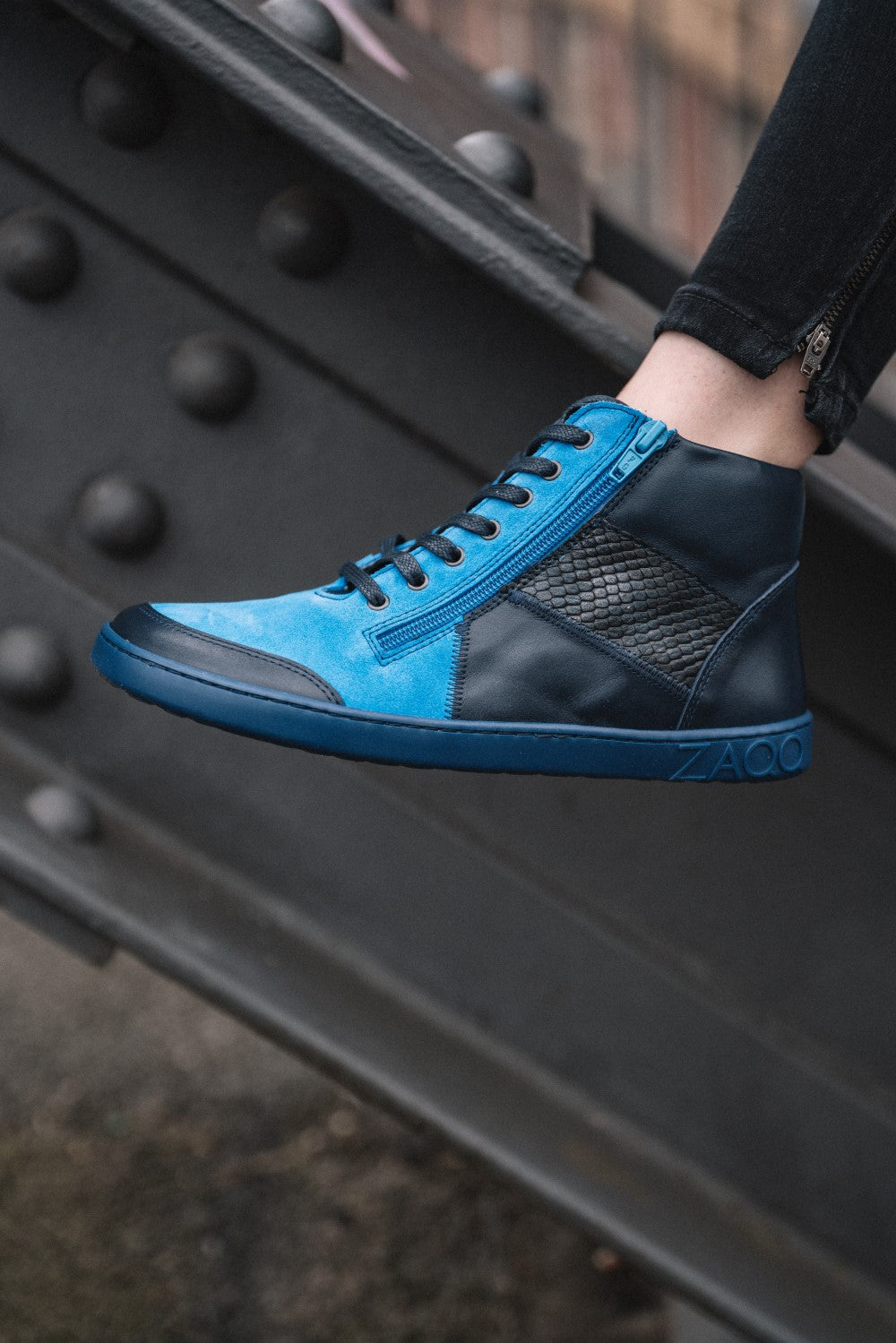 A close-up of a stylish azure and black high-top sneaker from LUQQ Azur collection ZAQQ, carried by a person standing on a metal support. The sneaker has a distinctive structural pattern, whereby the ZAQQ-Logo is featured prominently on the midsole. The person is wearing dark pants.