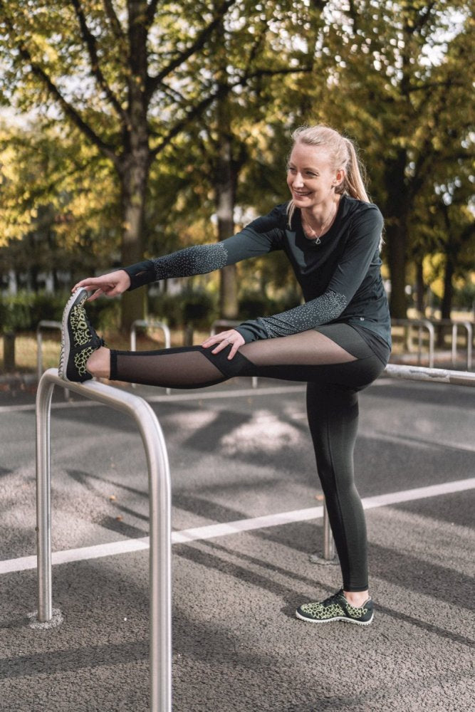 In the green sportswear QASEY from ZAQQ A woman stretches her legs outside on a metal railing. The breathable mesh outfit complements her bright smile as she is surrounded by trees with sunlight streaming through their leaves. The scene appears to be set in a park or outdoor training area.