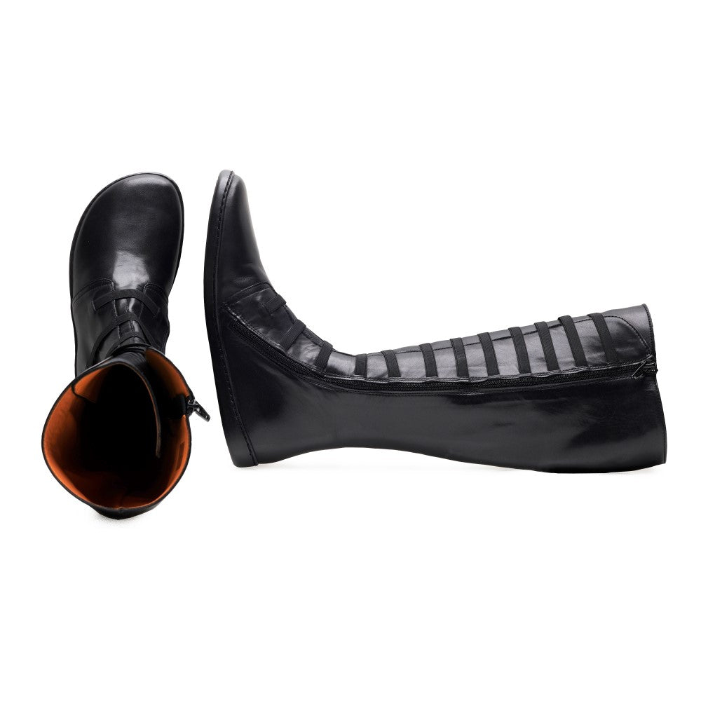 A pair of knee high gladiator boots QATCH Stretch Nappa Black from ZAQQ with open straps and flat rubber soles. One boot stands upright, highlighting the side zipper, while the other lies on its side, revealing the orange leather lining.