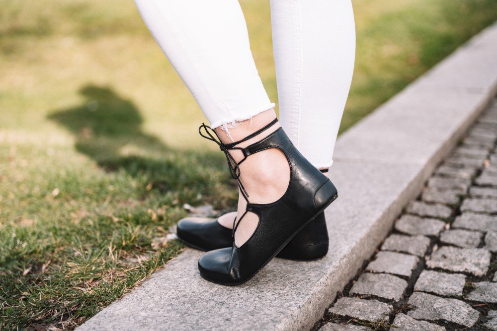 A person wearing black ballet flats and white pants ZAQQ carries, stands on a stone curb. The photo shows her feet and lower legs against a backdrop of grass and cobblestones, with shadows elegantly cast on the ground.