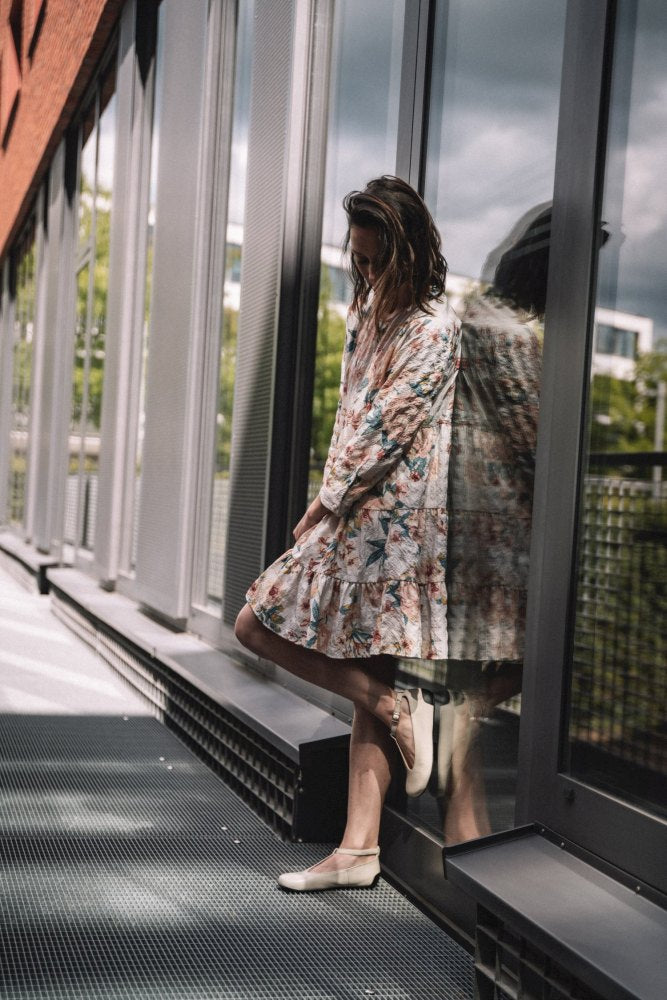 A person in a floral dress and QITE Beige ballerina flats ZAQQ leans against a reflective glass wall on an outdoor path. The scene is illuminated by natural light and complemented by greenery in the background.