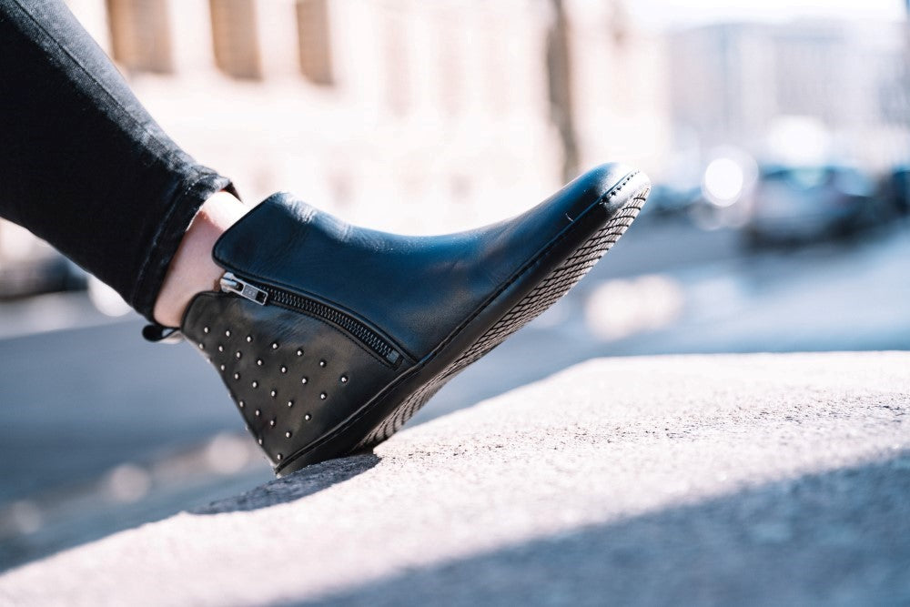 Close-up of a person wearing the black QLARE ankle boots ZAQQ wears, made of black leather with studded decorations and a side zipper, and stands on a concrete ledge. The non-slip sole provides stability against the blurred cityscape background illuminated by bright daylight.