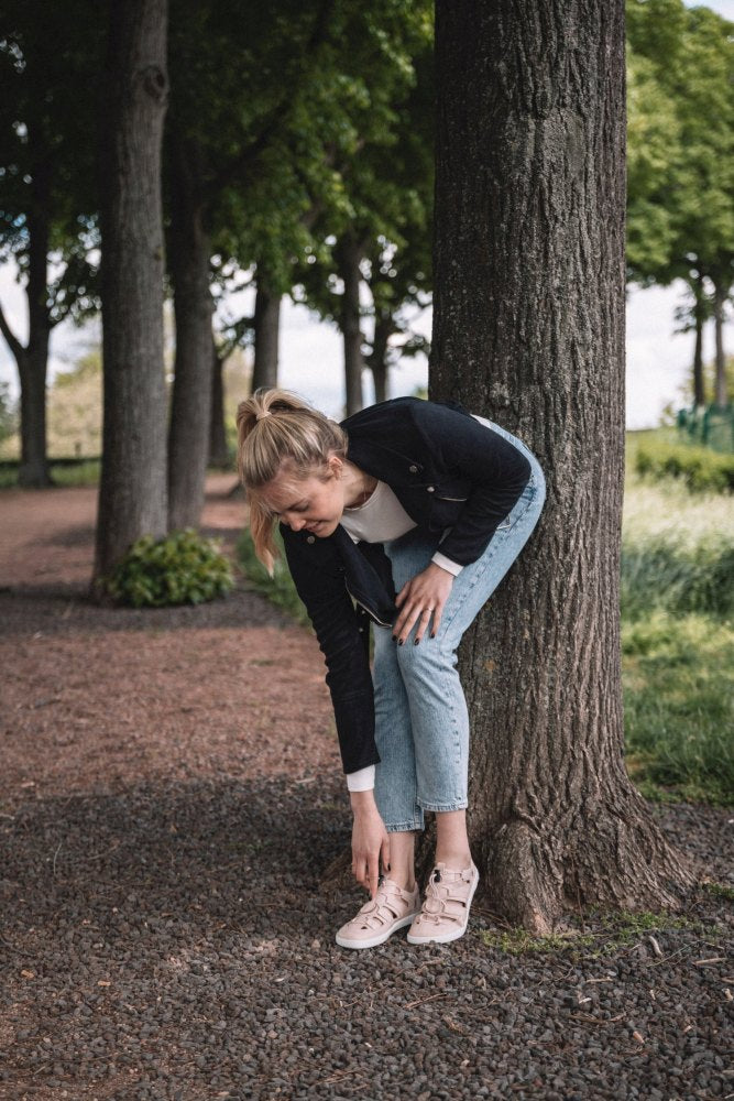 A woman with blonde hair, wearing a black jacket, a white shirt and jeans, bends down to pick up her QLEAR Light Pink ZAQQ as she leans against a tree on a tree-lined gravel path.