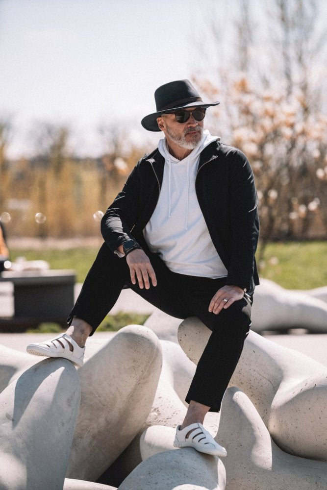 A person wearing a black hat, sunglasses, black jacket, white hoodie, black pants and chic QRUISE White sneakers from ZAQQ made of nappa leather sits outdoors on large stone sculptures. Trees and a blue sky can be seen in the background, adding to the tranquil atmosphere.