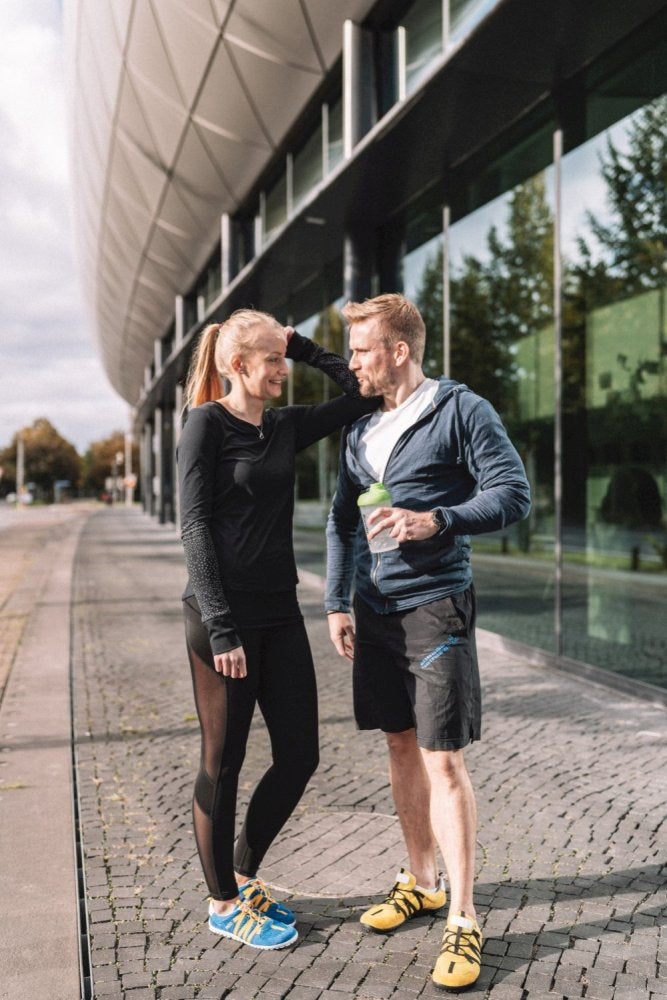 A man and a woman in sportswear stand in front of a modern building with glass windows. The woman, whose hair is tied back, smiles while the man holds a drink. Both seem relaxed in theirs ZAQQ RIQ Blue Orange shoes as they chat on the cobblestones.