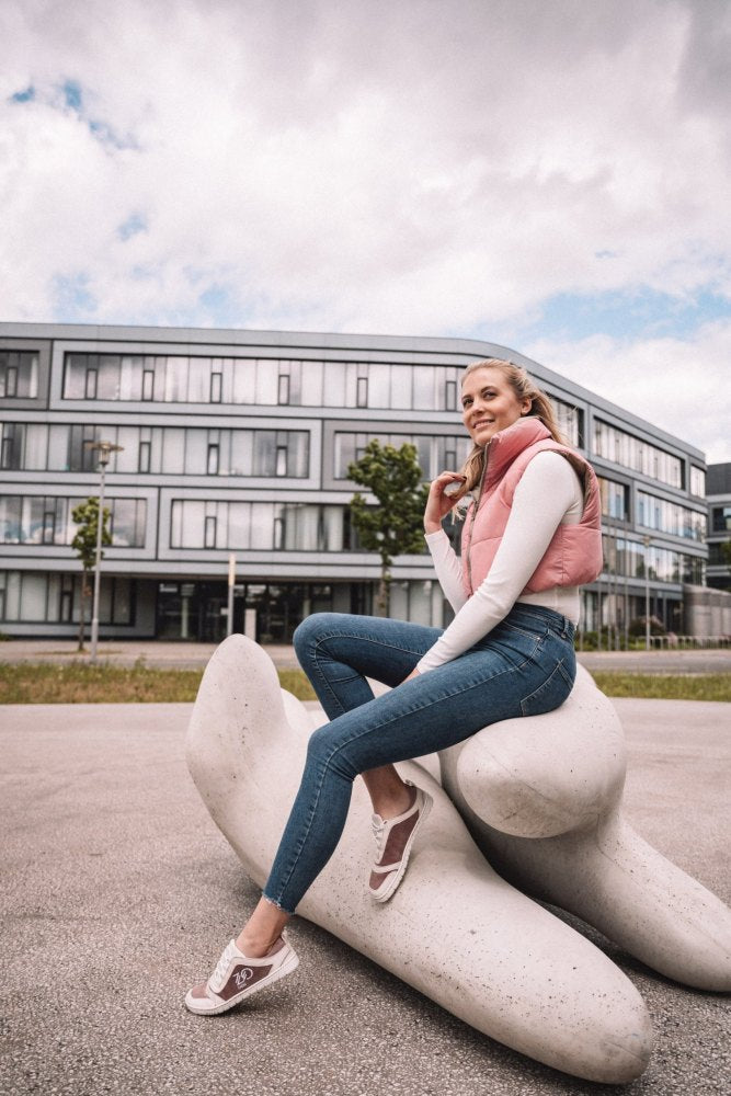 A person in a pink vest and jeans sits on a large abstract white sculpture in an urban setting and raises his stylish ZAQQ SQILL Rose vegan microfiber sneakers feature a modern building in the background and a cloudy sky above.