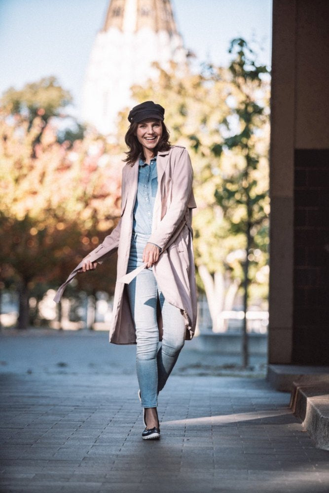 A woman confidently walks down a city street. She wears a long pink coat, a light blue jeans outfit and a black cap as well as chic dark blue shoes ZAQQ SUQAR. She smiles brightly, autumn trees and a blurred building can be seen in the background.