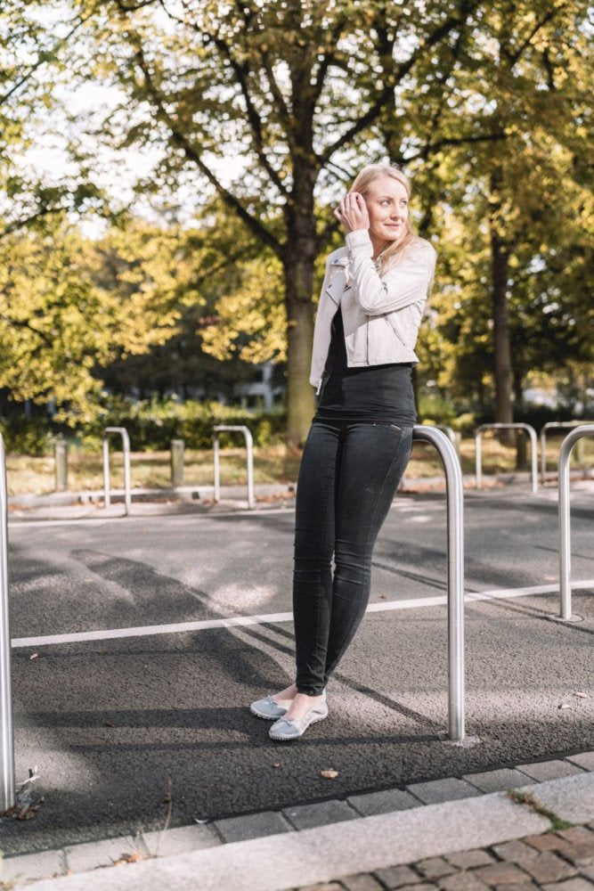 A person with long blonde hair, wearing a white jacket and dark jeans complemented by ZAQQ's SUQAR Gray shoes, stands on a street and leans on a metal post. Trees with green leaves can be seen in the background and sunlight shines through the branches.
