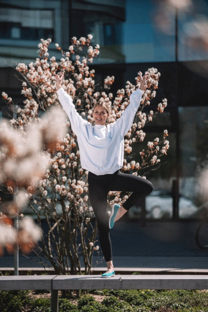 A person wearing a SUQAR Heaven sweater from ZAQQ and wearing black leggings, balanced on one leg next to a flowering tree, arms raised in a V, demonstrating perfect breathability. A modern building with glass windows can be seen in the background.