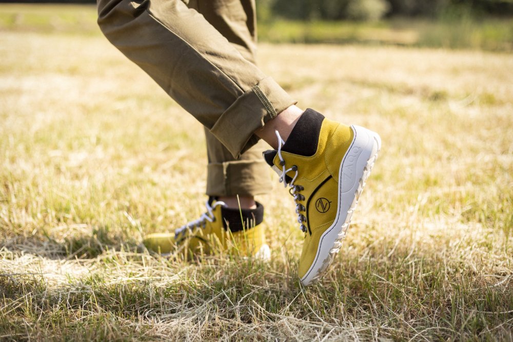 A person wearing TERRAQ Yellow Waterproof Sneakers from ZAQQ wearing, with high tops, white soles and dark laces, standing on dry grass. She wears rolled-up khakis against a sunny outdoor backdrop that perfectly showcases these sturdy outdoor shoes.