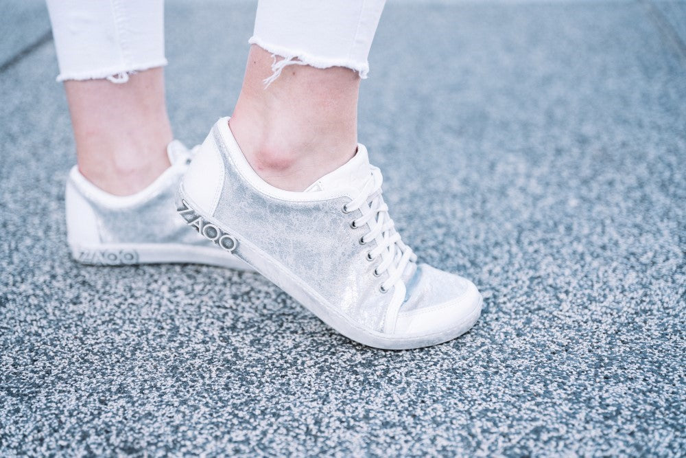 The feet stand on a textured gray-blue background, wearing a pair ZAQQ TIQQ Silver White sneakers made of premium nappa leather with white laces. The person is wearing slightly frayed white jeans.