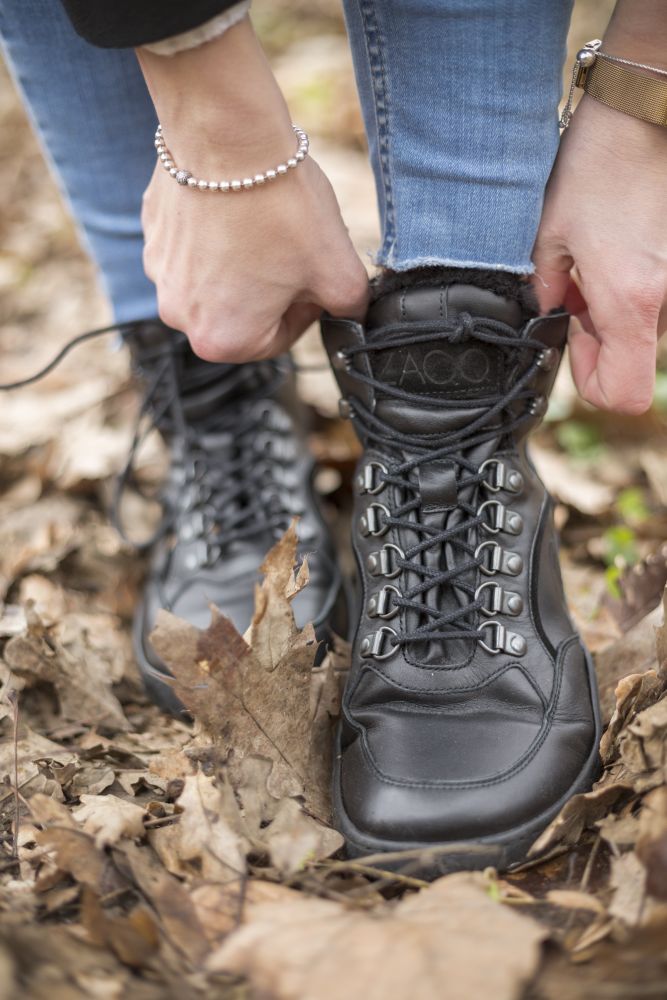A person in blue jeans ties the laces of their black TREQ waterproof winter boots ZAQQ, while standing on dry leaves. A bracelet adorns her wrist.