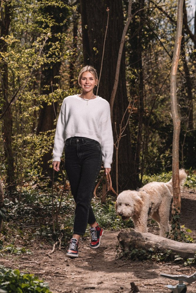 A woman in a white sweater and black pants walks a fluffy dog on a leash through a sunlit forest. With colorful VAQATION Red Waterproof Sneakers from ZAQQ she smiles as she strolls along the forest path surrounded by trees and greenery.