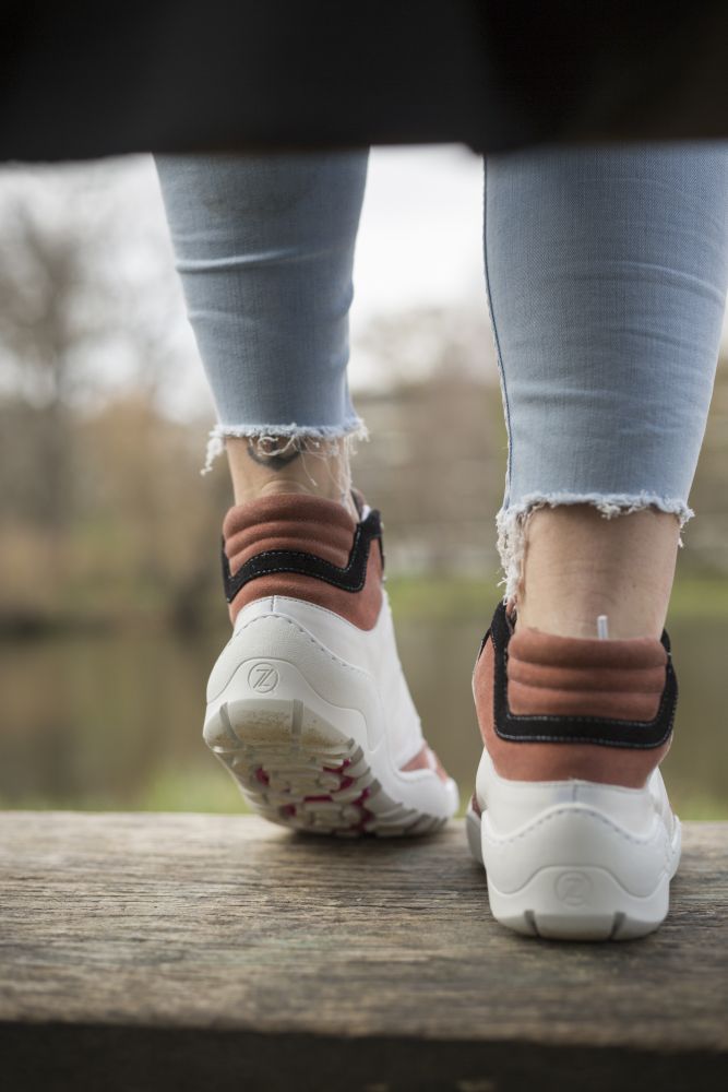 A person wearing frayed light blue jeans and white sneakers with pink and black accents stands on a wooden surface. The ZAQQ VAQATION Pink Waterproof Shoes ensure unparalleled breathability. A blurred view of a park with trees and a pond can be seen in the background.