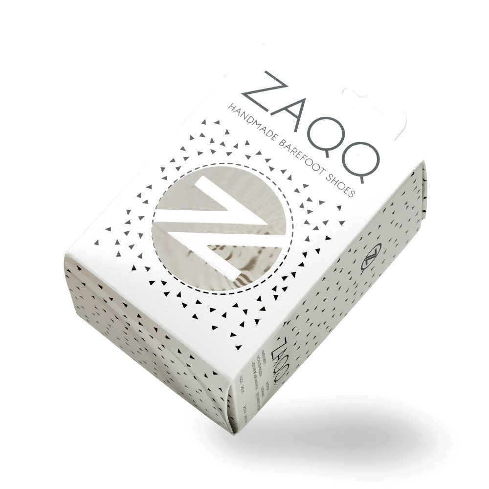 An oblique view of a white ZAQQ-Shoeboxes features geometric patterns and a round window that offers a sneak peek of the SOQQ Eclipse inside. The text reads “Handmade Barefoot Shoes”. The box with its subtle shadow sits gracefully on a plain background.