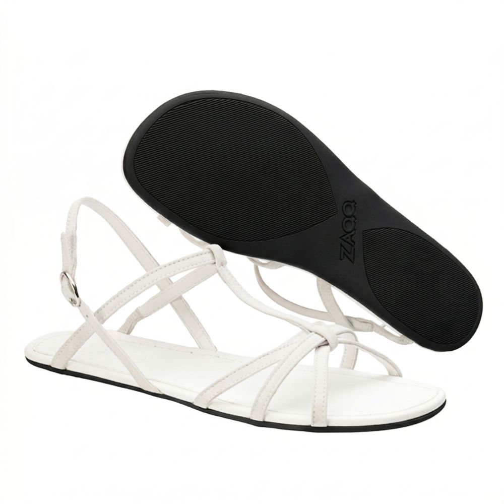 A pair of white strappy sandals ZAQQ QADA made of high-quality nappa leather with a flat sole. One sandal stands upright to reveal its elegant buckle, while the other is turned upside down to reveal the textured sole - the perfect mix of style and comfort.