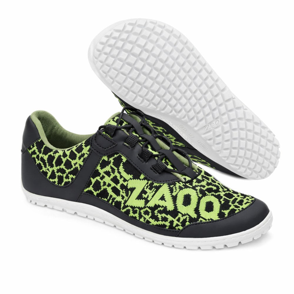 We present the QASEY Green from ZAQQ: a pair of casual shoes with an eye-catching black and green pattern design made from vegan materials. The shoes show that ZAQQ-Logo clearly visible on the side. They are equipped with a classic white sole with a barefoot shoe structure for better grip. One shoe is pictured standing upright while the other lies on its side to showcase the unique sole design.