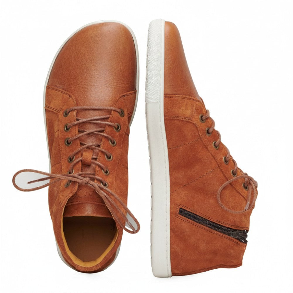 The QORE Mid Sierra from ZAQQ presents elegance as a pair of high-top sneakers in brown leather with a white sole. One sneaker is seen from above with the laces partially undone, while the other is viewed from the side and features a zipper detail and the brand name ZAQQ highlighted on the sole.