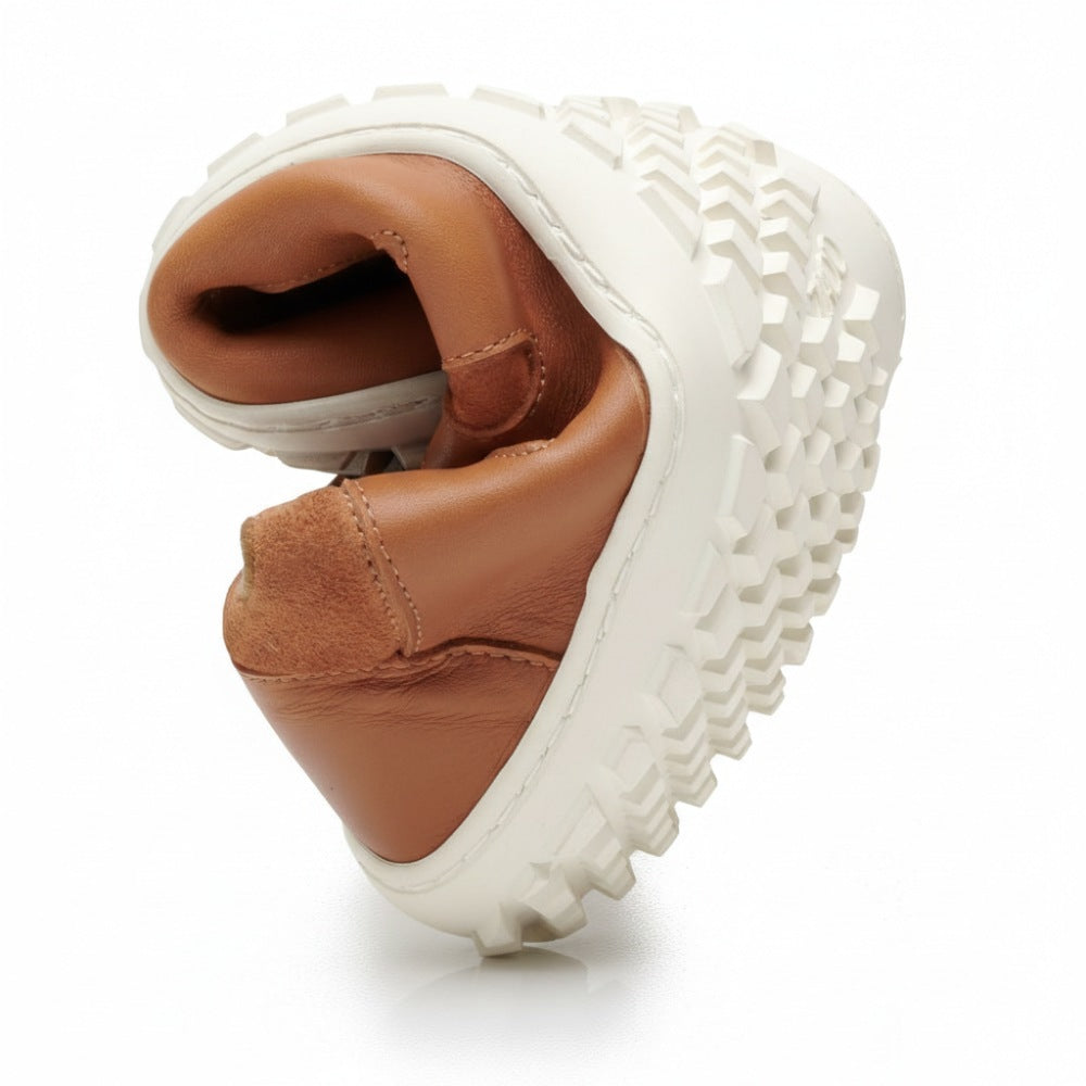 The SHOQQ cognac from ZAQQ, a tan leather sneaker with a thick white rubber sole, is folded in half to highlight its flexibility and comfort. The sole features a distinct, robust tread pattern similar to that of a barefoot shoe. The shoe stands against a simple white background.