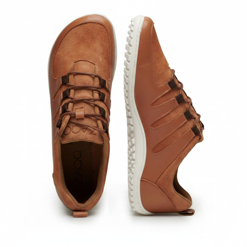 Two SHOQQ cognac sneakers from ZAQQ are presented. One is positioned upright to show the top view, while the other lies on its side and shows off the textured white sole design. These barefoot shoes have lace-up fronts and visible branding inside one shoe, highlighting their exceptional comfort.