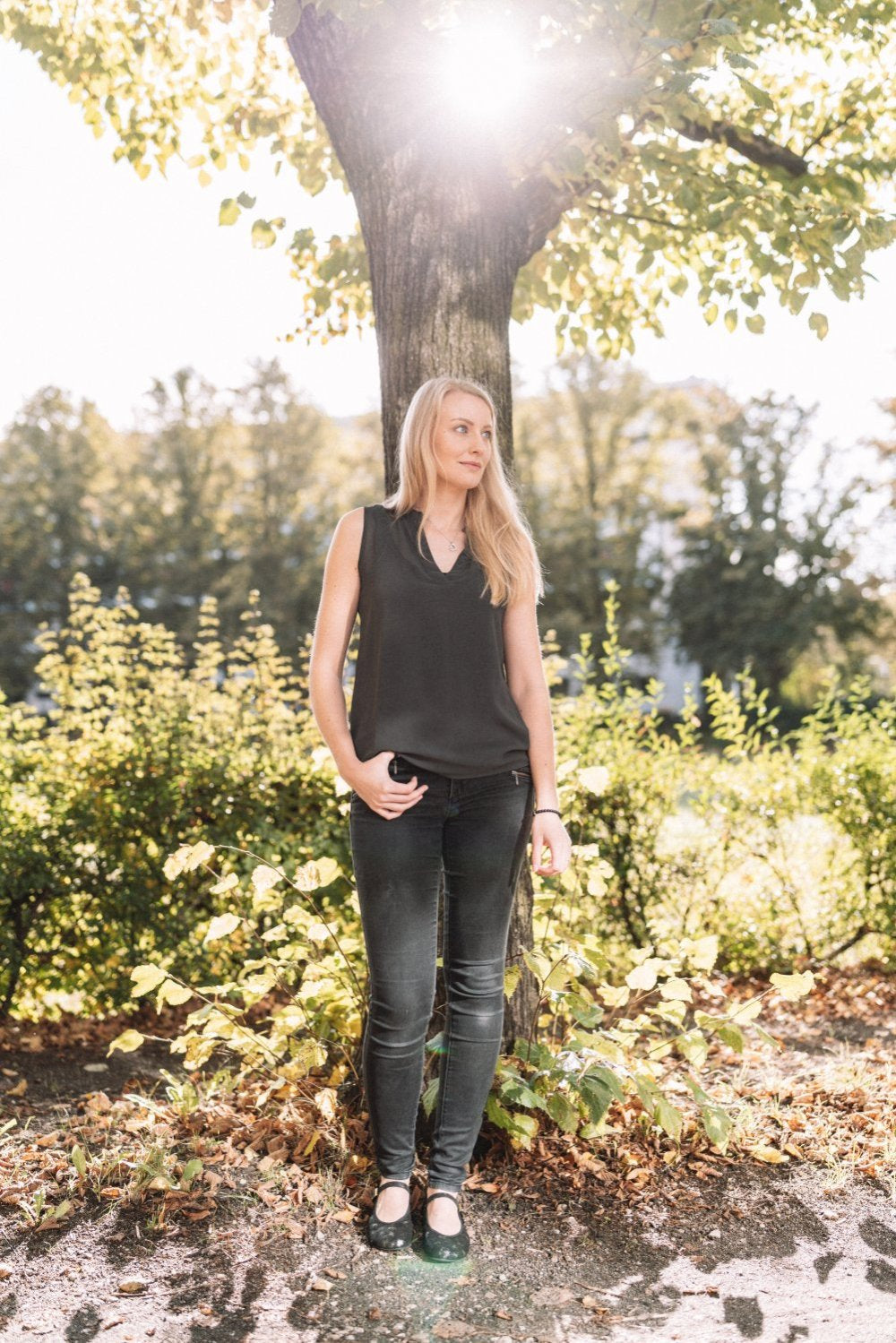 Eine Person mit langen blonden Haaren steht unter einem Baum in einem sonnenbeschienenen Park und strahlt lässige Eleganz aus. Sie trägt ein ärmelloses schwarzes Top und Jeans, eine Hand in der Tasche und hat einen entspannten Gesichtsausdruck, während das Licht durch die Blätter darüber fällt. Ihre TWIST Ennis-Schuhe von ZAQQ ergänzen die ruhige Umgebung.